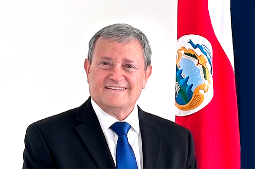 Retrato oficial de Guillermo Carazo Ramírez, Ministro, sonriendo con traje formal oscuro frente a la bandera de Costa Rica